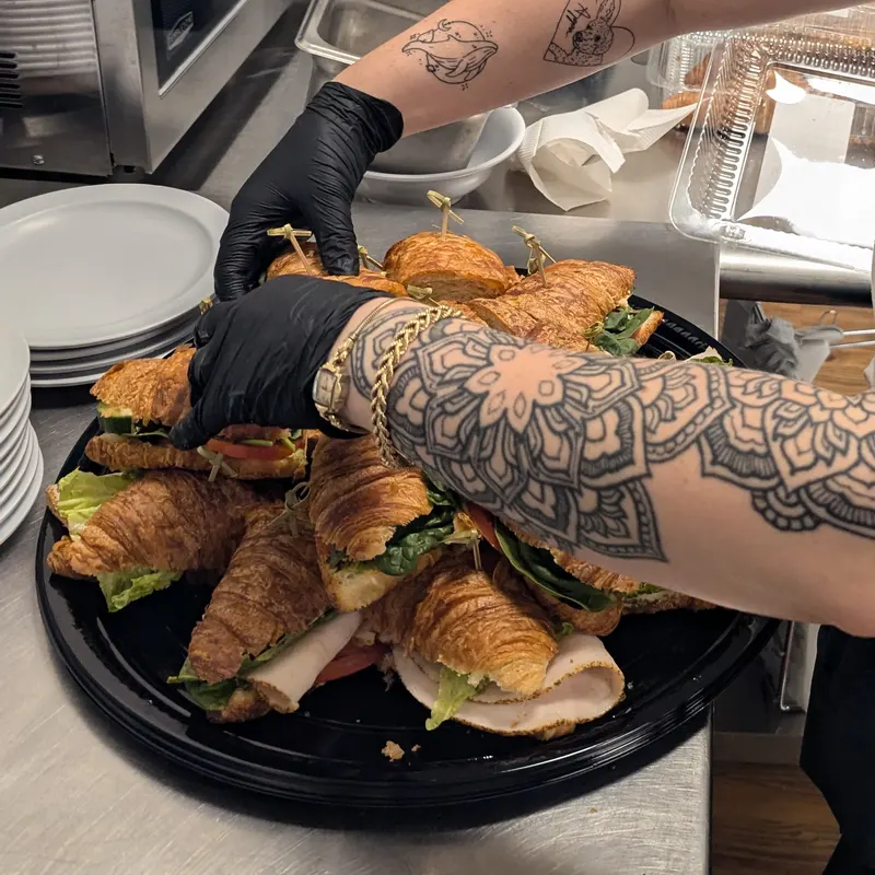 Gloved hands arranging halved croissant sandwiches with turkey and lettuce on a round black catering platter.