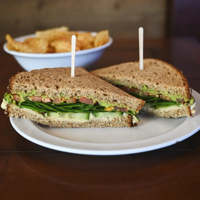 The Rustic cut in half on seeded bread, layered with spinach, cucumber and avocado spread, with a bowl of chips behind it.
