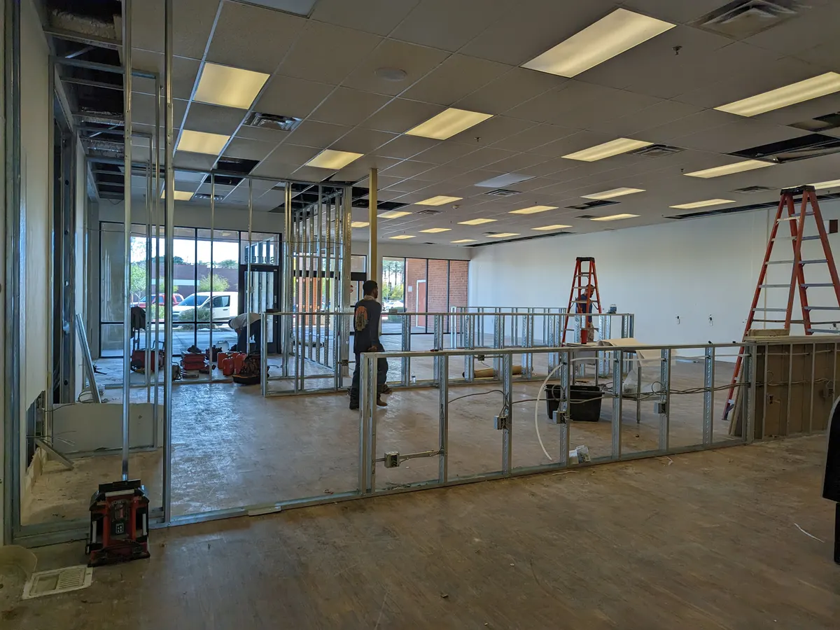 The empty unit during the build-out: metal stud walls, a bare floor, ladders, and a worker in the middle distance.