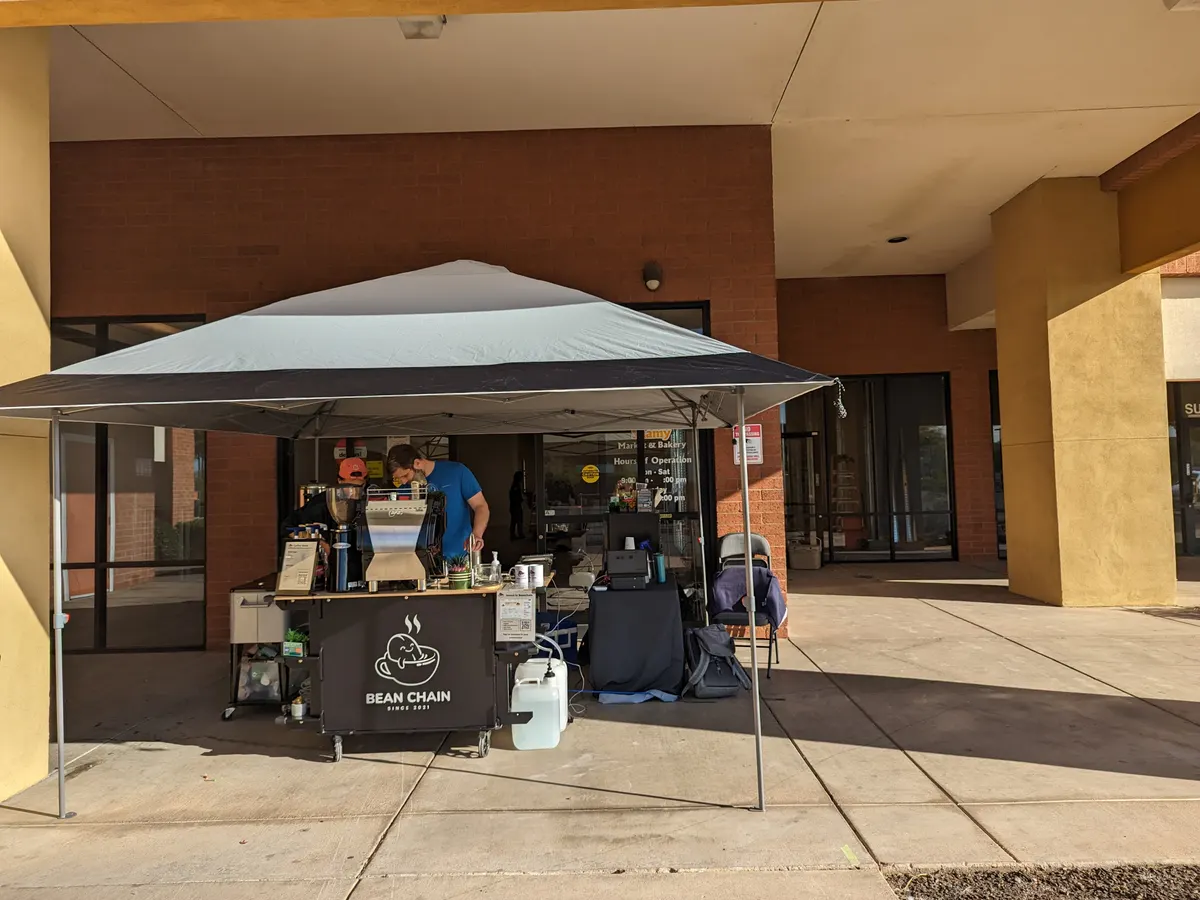 The Bean Chain coffee cart under a white pop-up canopy on the sidewalk outside the plaza, two people at the espresso machine, and ladders visible through the glass beside it.