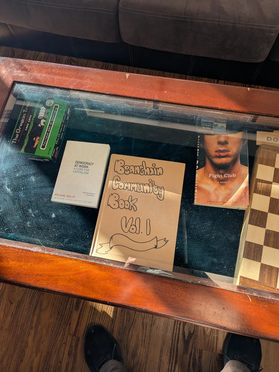 The Beanchain Community Book, volume one, hand lettered on a kraft cover, lying under glass beside a copy of Democracy at Work by Richard Wolff and a chessboard.