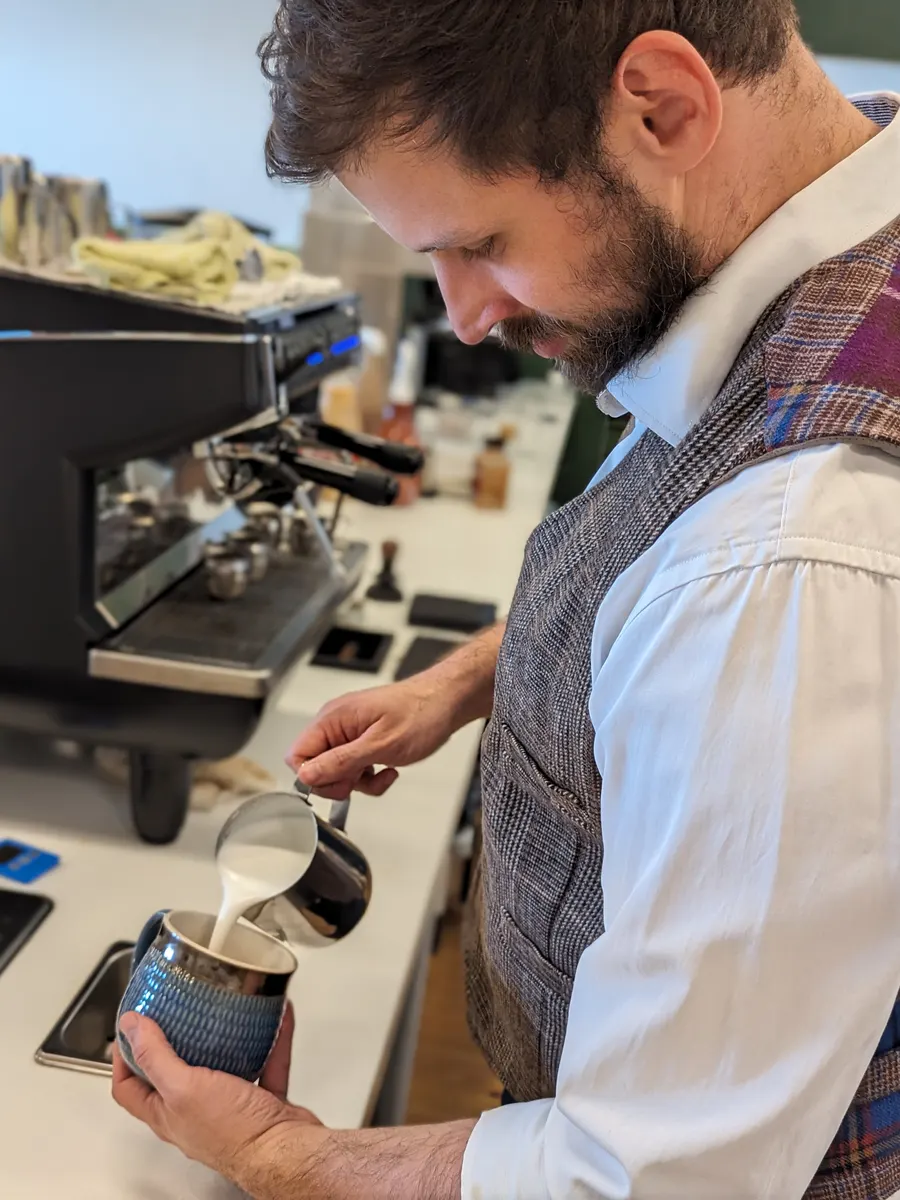 A barista in a tweed vest pouring steamed milk into a blue ceramic mug beside the espresso machine.