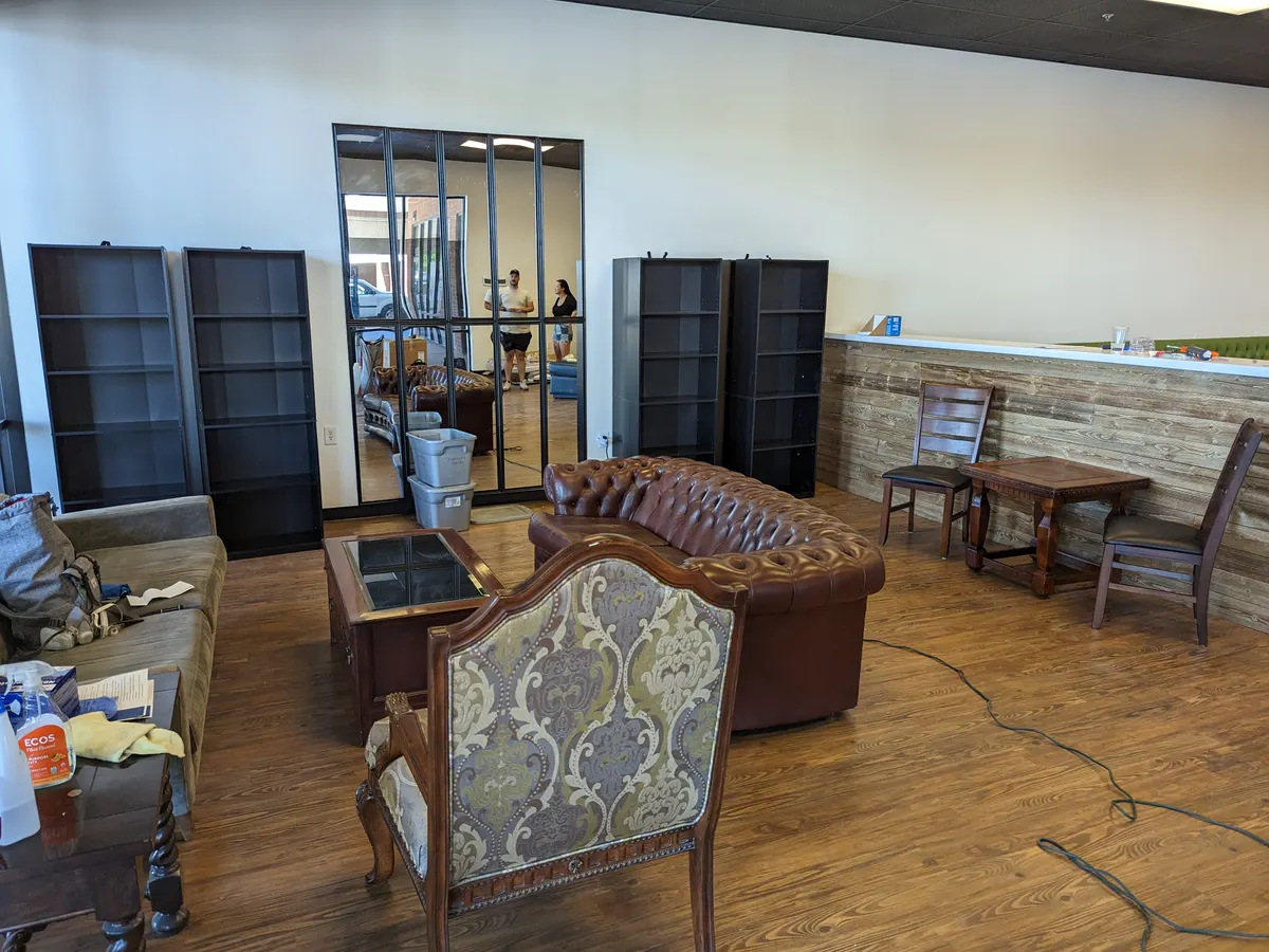 The corner that became the library: empty black bookshelves, a brown leather chesterfield, an upholstered armchair and a small wooden table.