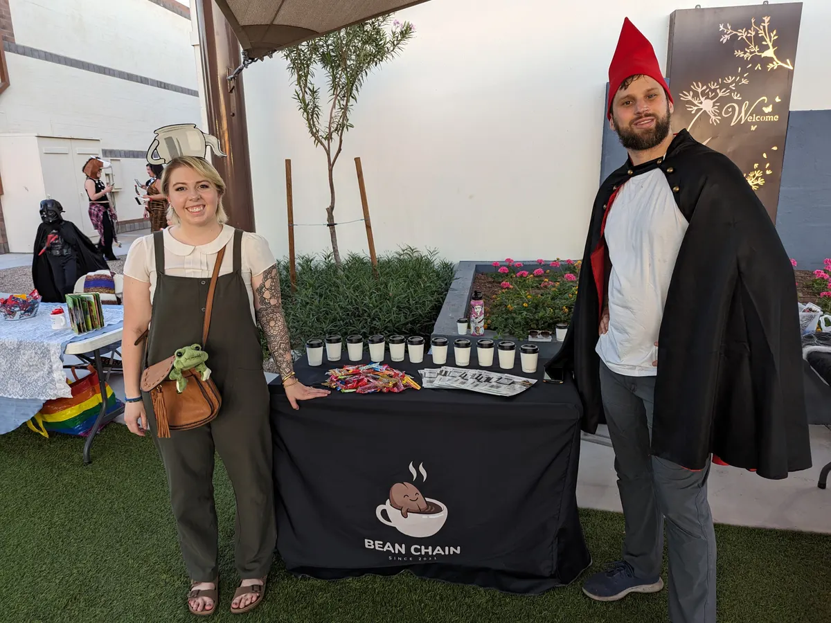 Two people in costume, one in a teapot hat and one in a red pointed hat and black cape, beside a Bean Chain table of coffee cups and sweets at an outdoor event.