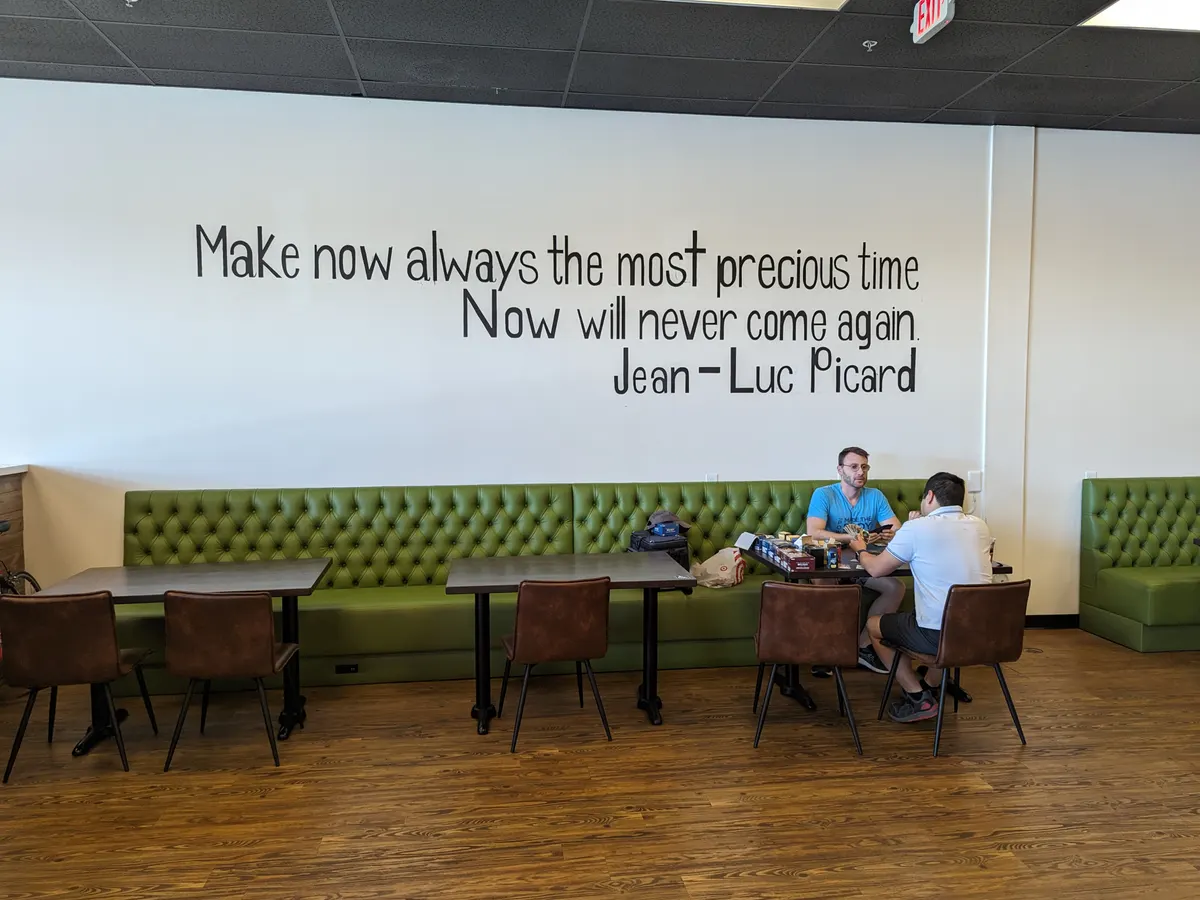 A quotation painted on the white wall above the green banquette, reading: Make now always the most precious time. Now will never come again. Jean-Luc Picard. Two people talk at a table below it.