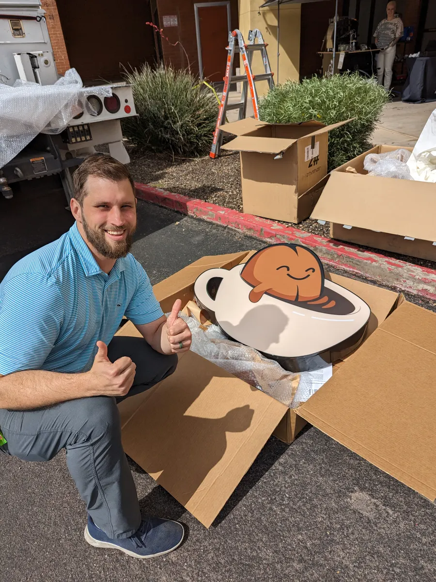 Somebody crouching beside an open cardboard box in the parking lot, giving a thumbs up next to the new sign: the smiling bean sitting in a coffee cup.
