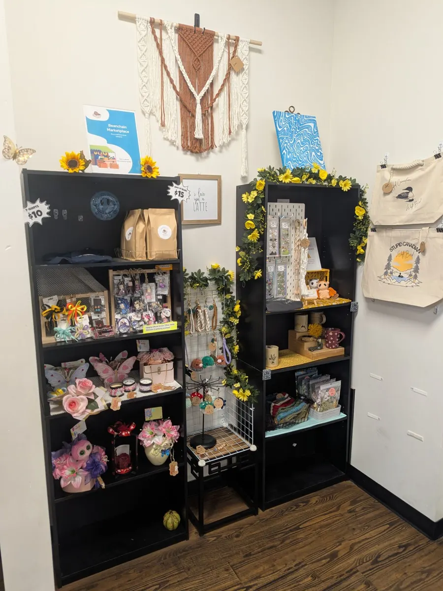 The vendor shelves at Beanchain: two black bookcases of crochet animals, candles, flowers, jewelry and prints, with a macramé hanging and tote bags on the wall beside them.