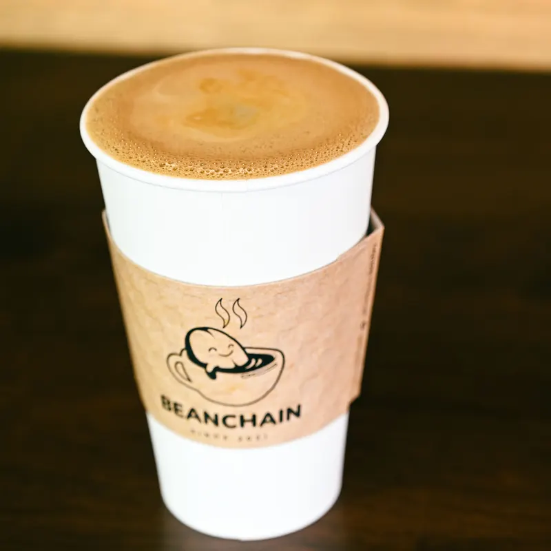 A hot drink in a Beanchain to-go cup with the bean and cup logo on the sleeve.
